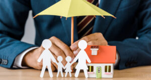Image of paper cutout of family in front of home model with umbrella over house and cutout family on desk of insurance agent